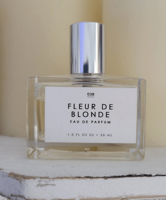 Gourmand Eau de Parfum in Fleur De Blonde sitting on a white dresser in a clear bottle with a long silver cap.