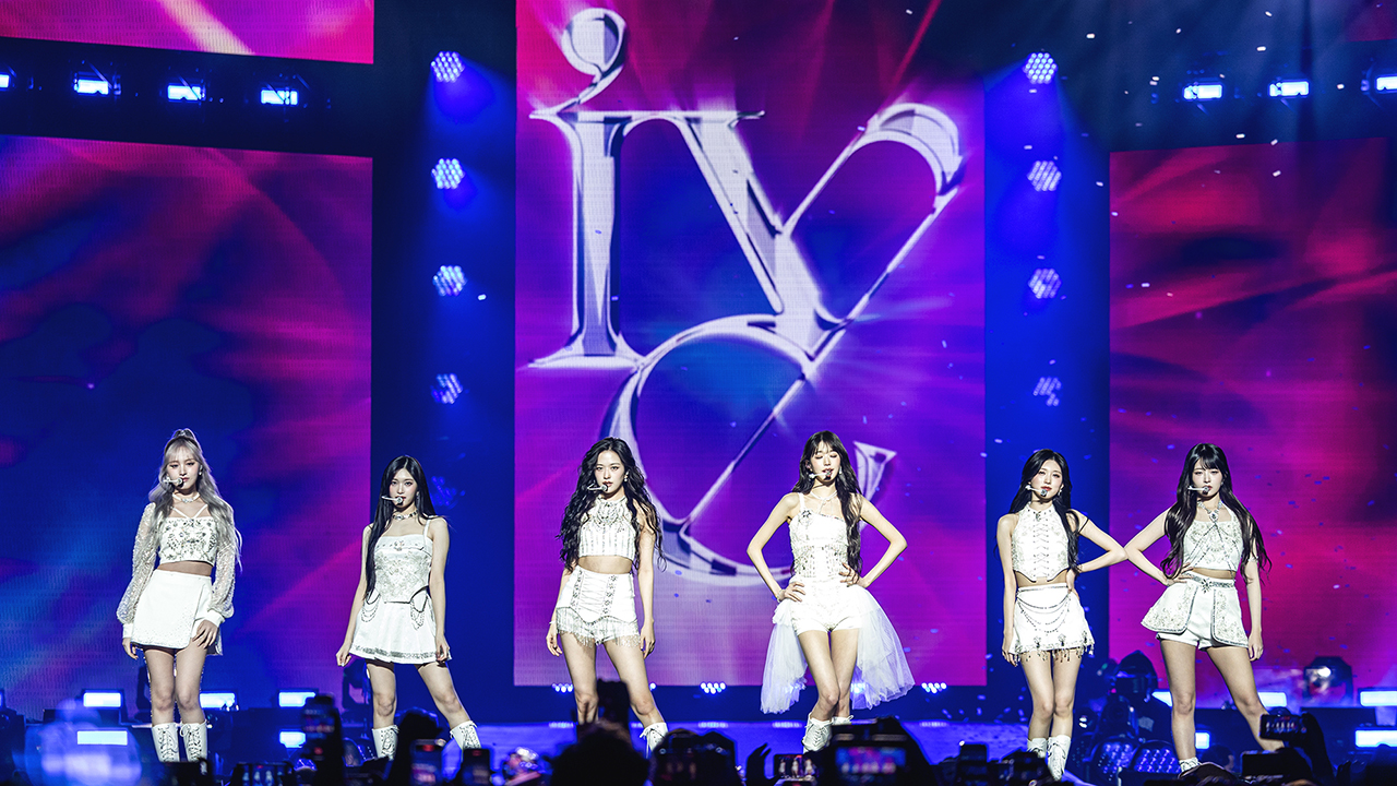 K-Pop Girl Group IVE, six Asian women ANYUJIN, GAEUL, REI, JANGWONYOUNG, LIZ, and LEESEO, standing with white outfits on stage.