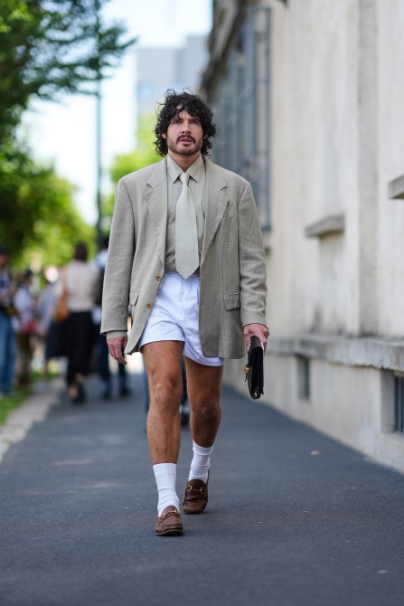 Luke Day wears a oversized gray green blazer jacket, gray green button up long sleeve, white short shorts, black clutch, white long socks, brown leather loafers, outside Prada, during the Milan Fashion Week Menswear Spring/Summer 2025 on June 16, 2024 in Milan, Italy.
