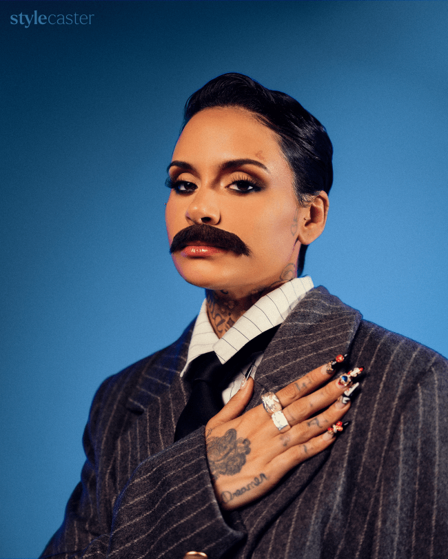 Kehlani wearing a suit and mustache holding her hand over heart