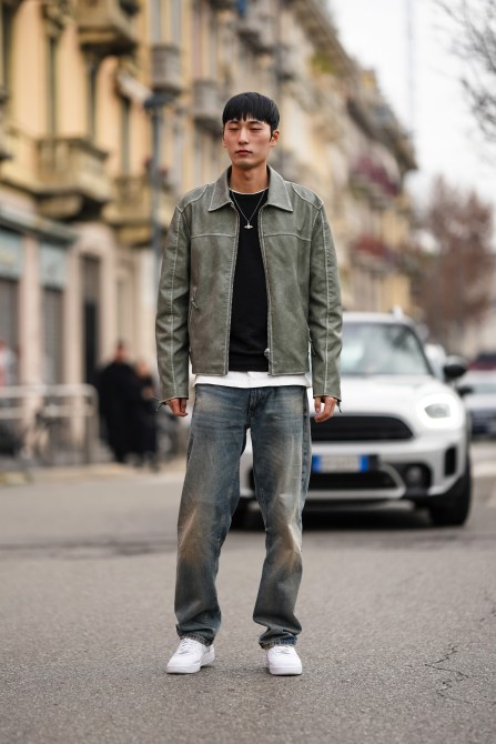 A man wears a necklace , a bb pullover , a gray leather jacket , a white long t-shirt , denim pants / jeans , white sneakers shoes,