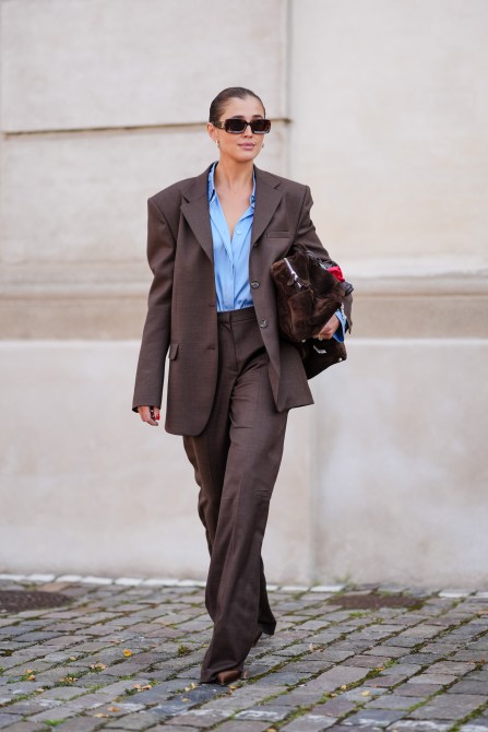 Darja Barannik wears dark brown sunglasses, silver earrings, light blue button up long sleeve shirt, dark brown oversized blazer jacket dark brown loose pants, dark brown suede bag, mixed metal rings, dark brown shoes, outside The Garment, during the Copenhagen Fashion Week Spring/Summer 2024-2025 on August 7, 2024 in Copenhagen, Denmark.
