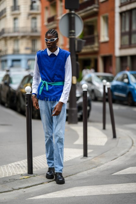 A guest wears white sunglasses from Marine Serre, a white shirt, a royal blue sleeveless / fringed pullover, blue faded denim large pants, black shiny leather laces ankle boots, a blue tie as a belt, outside the Dior Homme show, during Paris Fashion Week - Menswear Spring/Summer 2023, on June 24, 2022 in Paris, France.