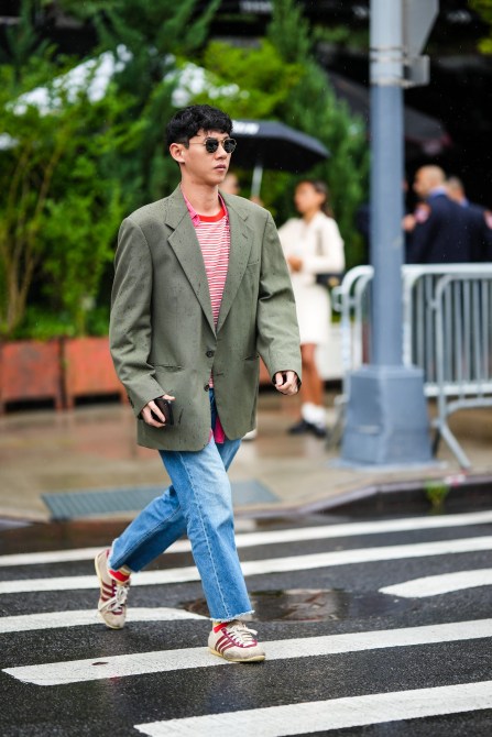 A guest wears black circle sunglasses, a red and beige striped print pattern t-shirt, a red long shirt, a khaki blazer jacket, blue denim faded ripped jeans pants, beige suede and burgundy shiny varnished leather logo sneakers from Adidas, outside Khaite, during New York Fashion Week, on September 11, 2022 in New York City.