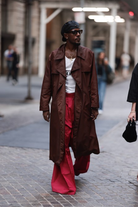 A guest seen wearing Prada black nylon logo hat, brown sunglasses, diamond stud earrings, gold cross pendant necklace, white cotton tank top, brown nylon long coat, red long flared trousers, before the WILLY CHAVARRIA show, on September 06, during the New York Fashion Week, 2024 in New York City.