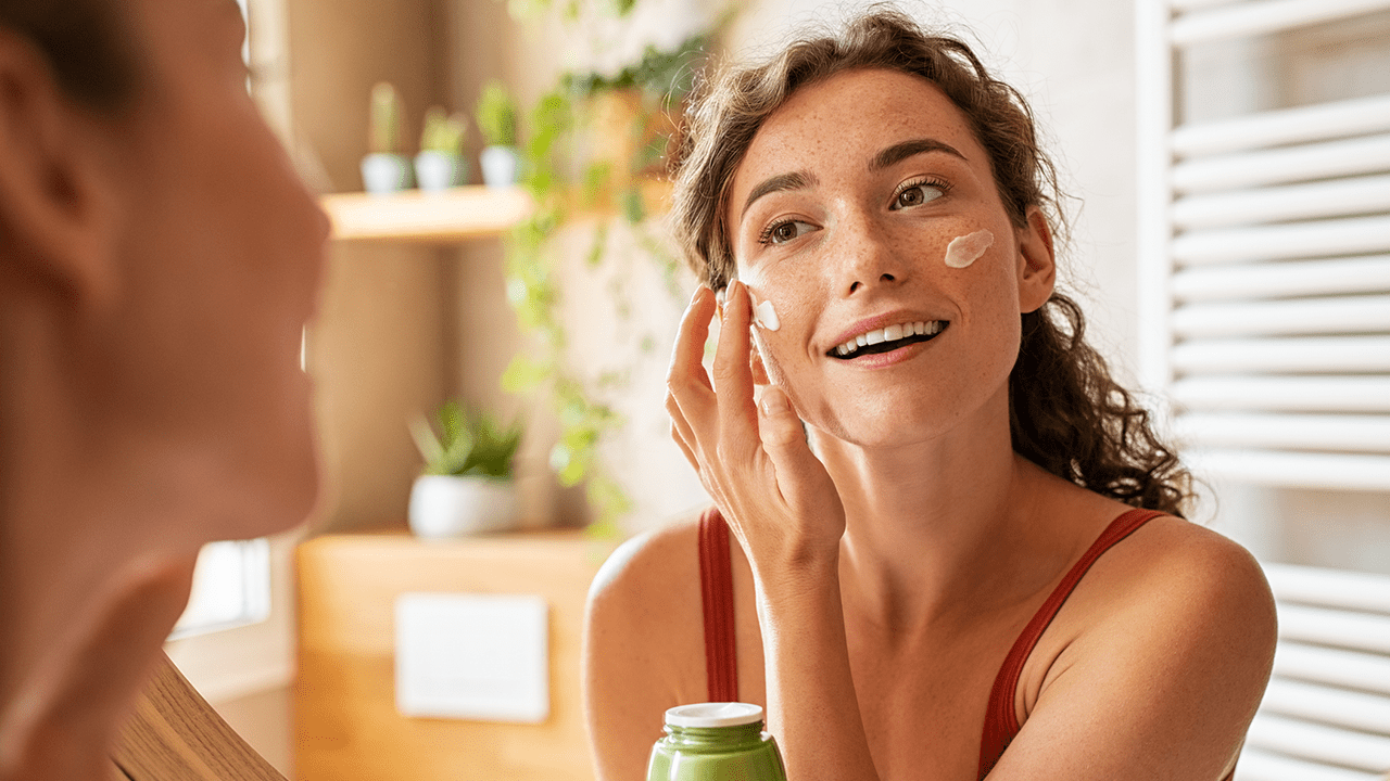 Woman caring of her beautiful skin face standing near mirror in the bathroom. Young woman applying moisturizing cream on her face during morning routine. Smiling natural girl holding little green jar of ecological cosmetic cream.